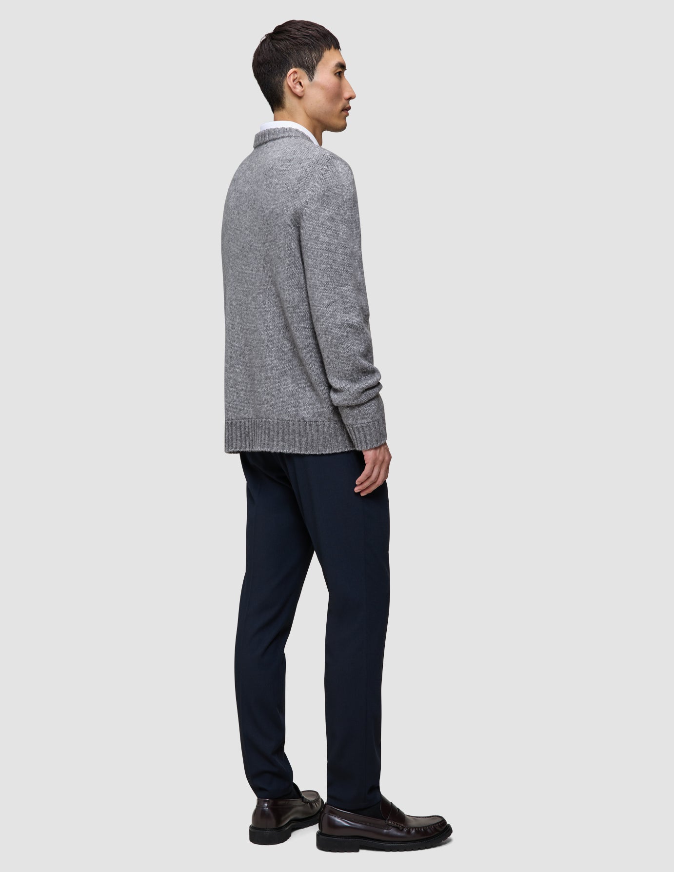 En man, sedd i profil, bär en tjock, mjuk ulltröja med rund hals i färgen Glacier Grey över en skjorta med krage, mörka byxor och loafers.